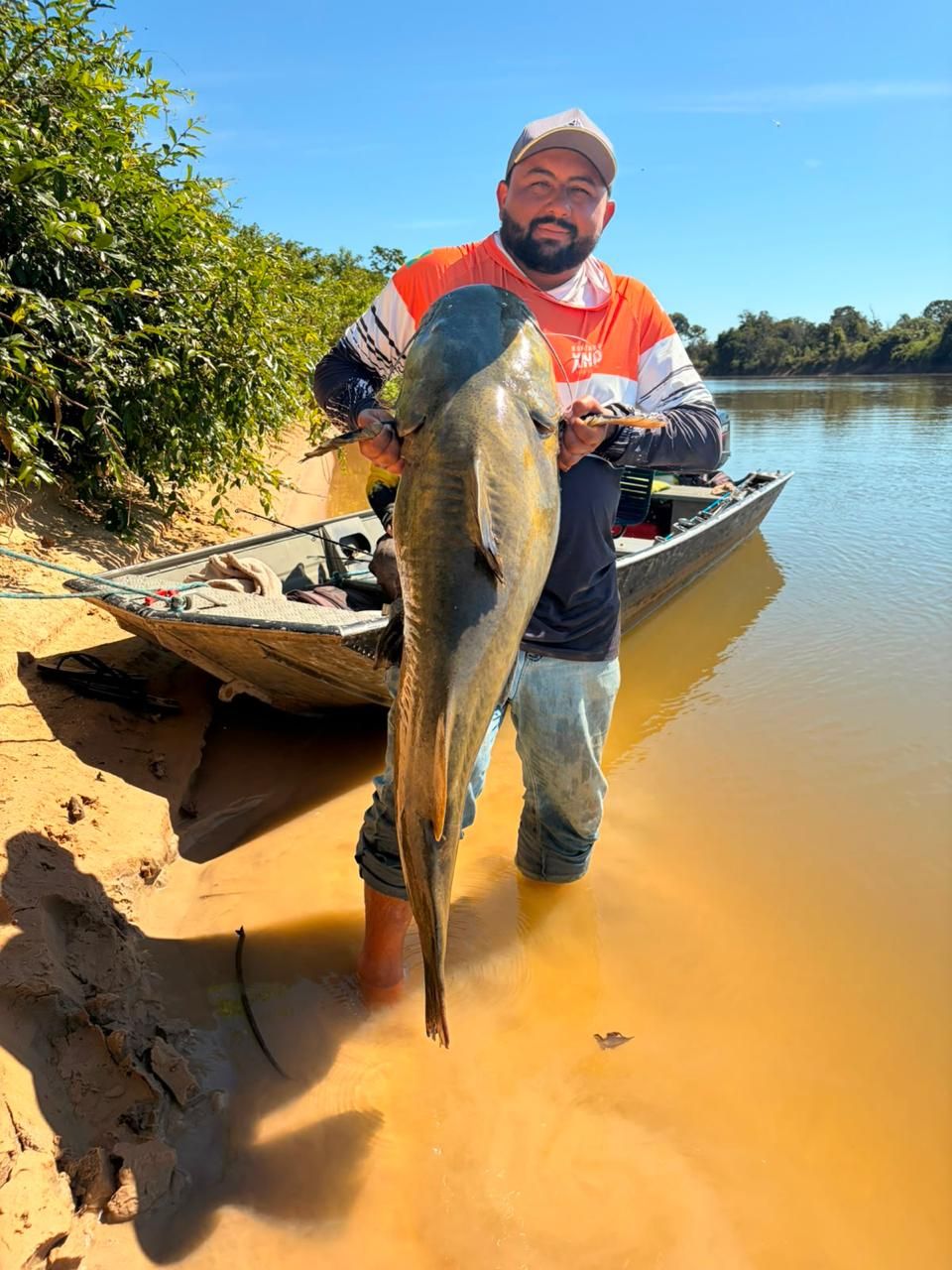 Angler standing in amber shallows holding a catfish beside a boat